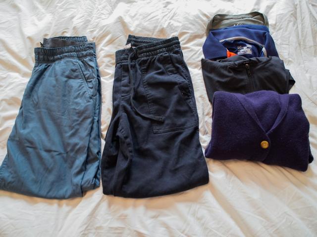 From left to right: Folded teal pants, folded black pants, and a row of two rolled-up shirts and two fold-up long sleeve layers that are gray, black, and navy blue on a white sheet
