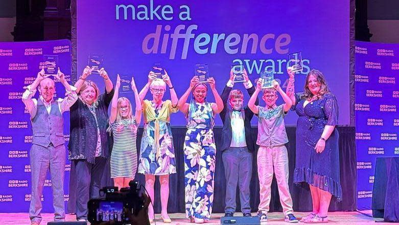 Eight winners stand in a line on a stage with purple backdrop holding their wards above their heads