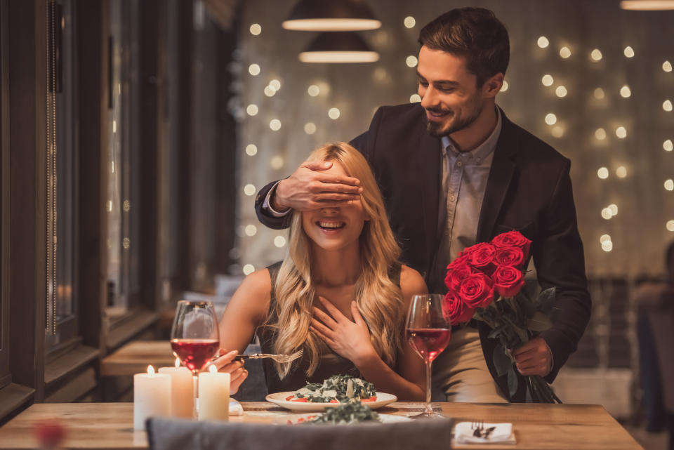 Handsome elegant man is holding roses and covering his girlfriend's eyes while making a surprise in restaurant, both are smiling