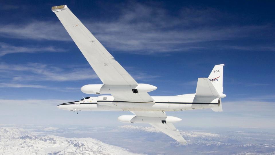 a white aircraft with a large wingspan flies above snowcapped mountains and deserts