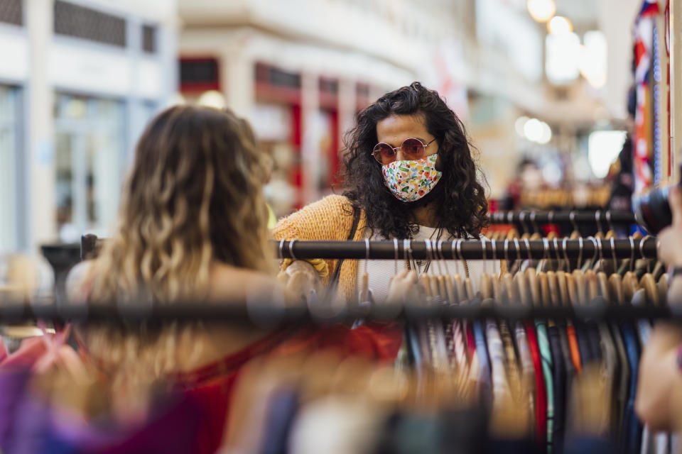 A shot of a young caucasian hipster woman and her mid adult Pakistani boyfriend looking at second hand, recycled clothing at a small local business in a market. They are wearing casual bohemian clothing, accessories, eyewear and reusable protective face masks.