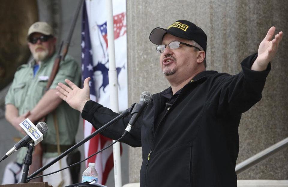 A man wearing a black shirt and baseball cap speaks to a crowd via a microphone