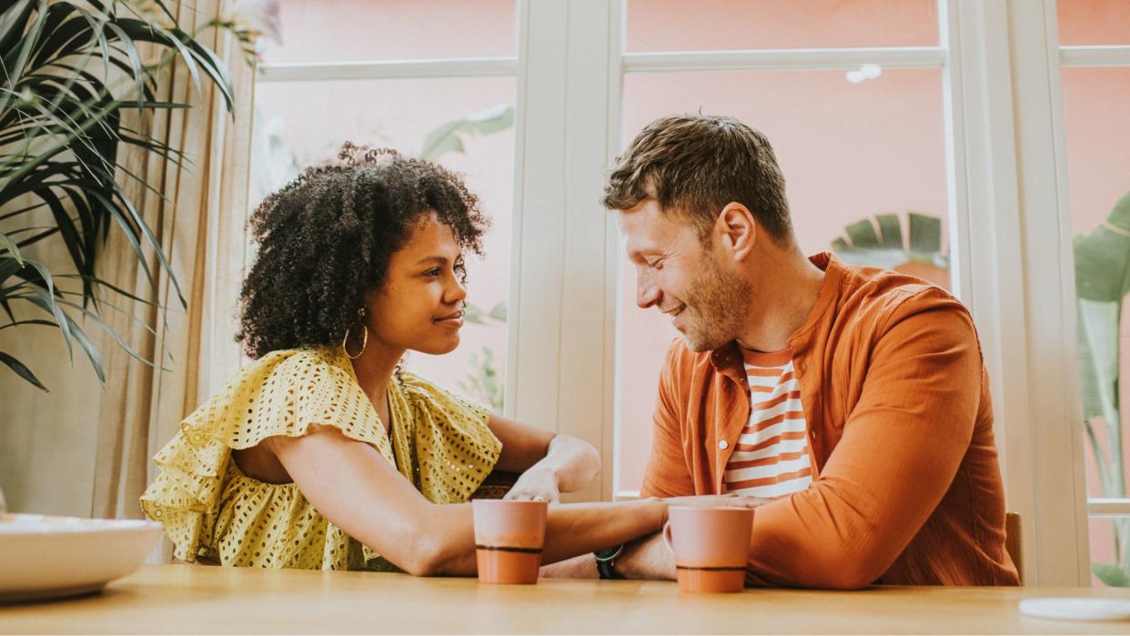  Woman and man sit next to each other at a coffee shop, smiling softly as if on a date. 