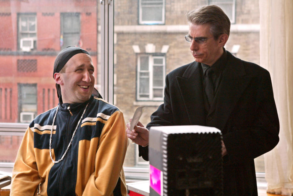 Richard Belzer in a black suit and sunglasses hands a document to a man in casual sportswear with a smile, standing near a window with a cityscape view