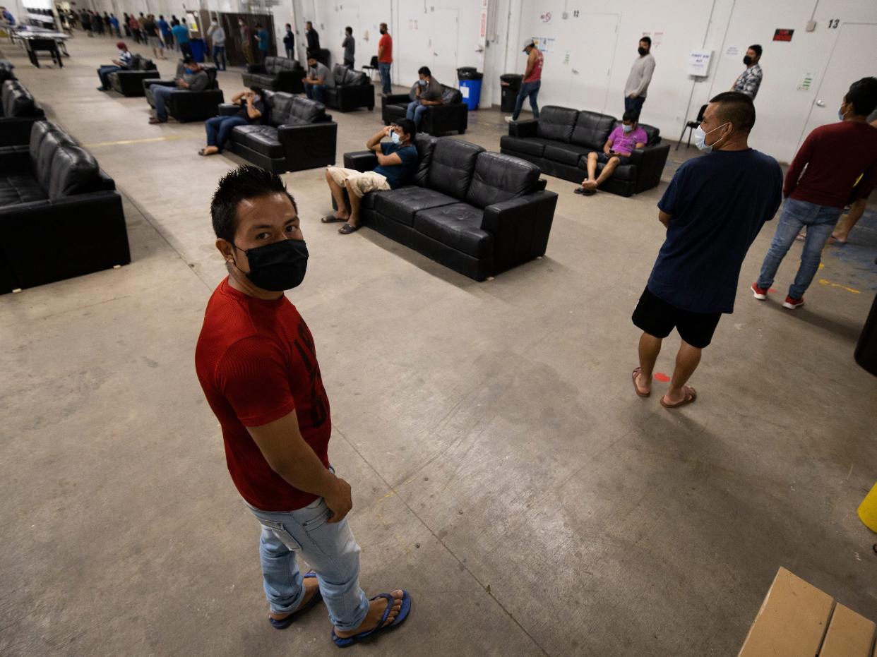 A migrant farm laborer in a face mask is in line for food in a living space filled with black couches.