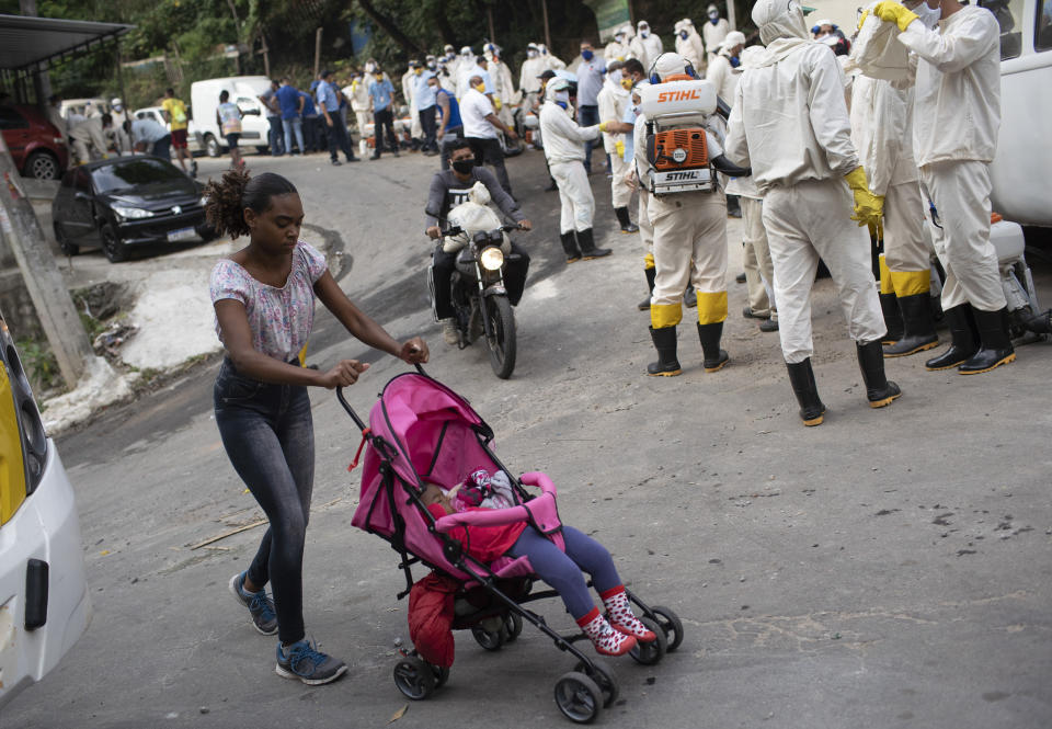 Una mujer que empuja un cochecito de bebé pasa junto a los trabajadores de servicios públicos de agua que desinfectarán la favela de Turano en un esfuerzo por frenar la propagación del nuevo coronavirus en Río de Janeiro, el martes 9 de junio de 2020. (AP Foto/Silvia Izquierdo)