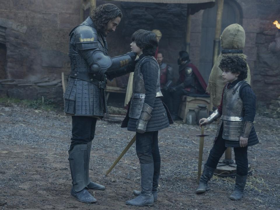 A man wearing armor and standing in front of a younger boy who is training with a wooden sword.