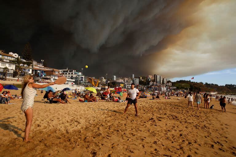 Turistas juegan a las palas en una playa con una gran nube de humo negro a sus espaldas, procedente de los incendios forestales, en Viña del Mar, Chile, el viernes 2 de febrero de 2024. (Martín Thomas, Aton Chile vía AP)