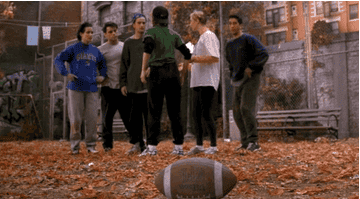 A group of people rush to grab a football on a leafy field