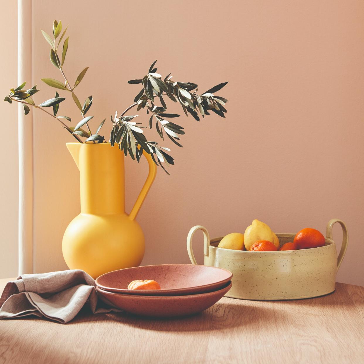  A wooden dining table against a pink wall with a yellow jug and and a bowl of citrus fruit on top. 