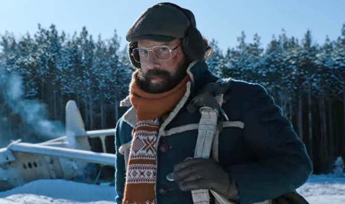 Murray wears a hat, gloves, scarf, and warm coat as he stands in front of a broken-down plane near the snowy forest