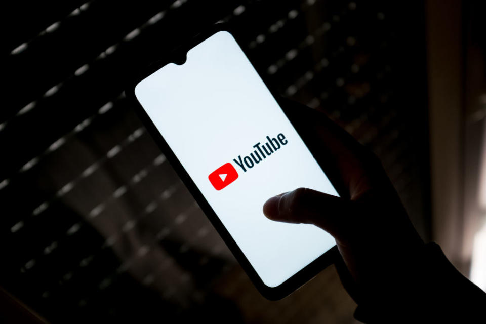 Hand holding a smartphone displaying the YouTube logo onscreen, backlit by window blinds