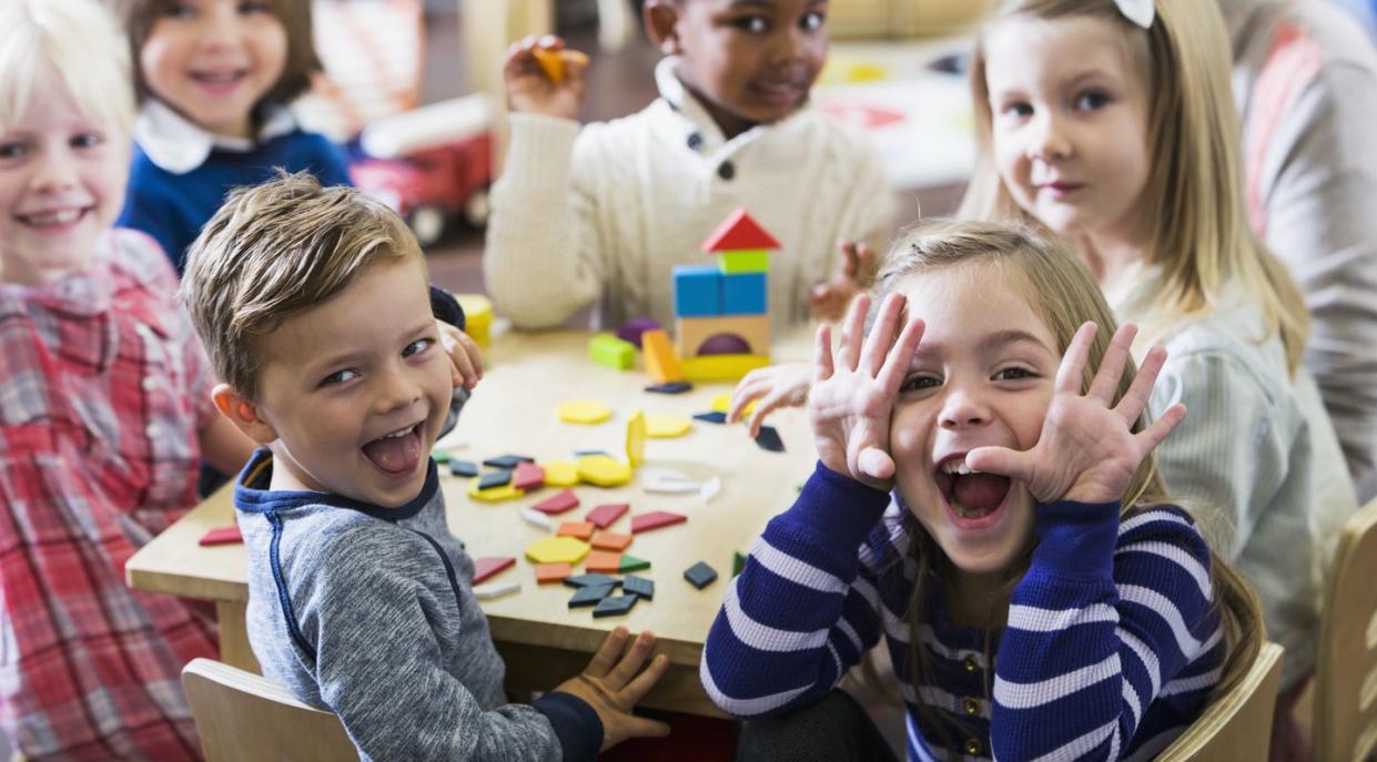 a multiracial group of preschoolers or kindergarteners having fun in the classroom six children are sitting around a little wooden table playing with colorful wooden block and geometric shapes the playful little girl in the foreground is making a silly face at the camera
