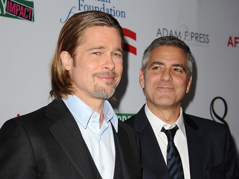 Brad smiling in a three-piece suit and George looking at him and smiling in a suit and tie.