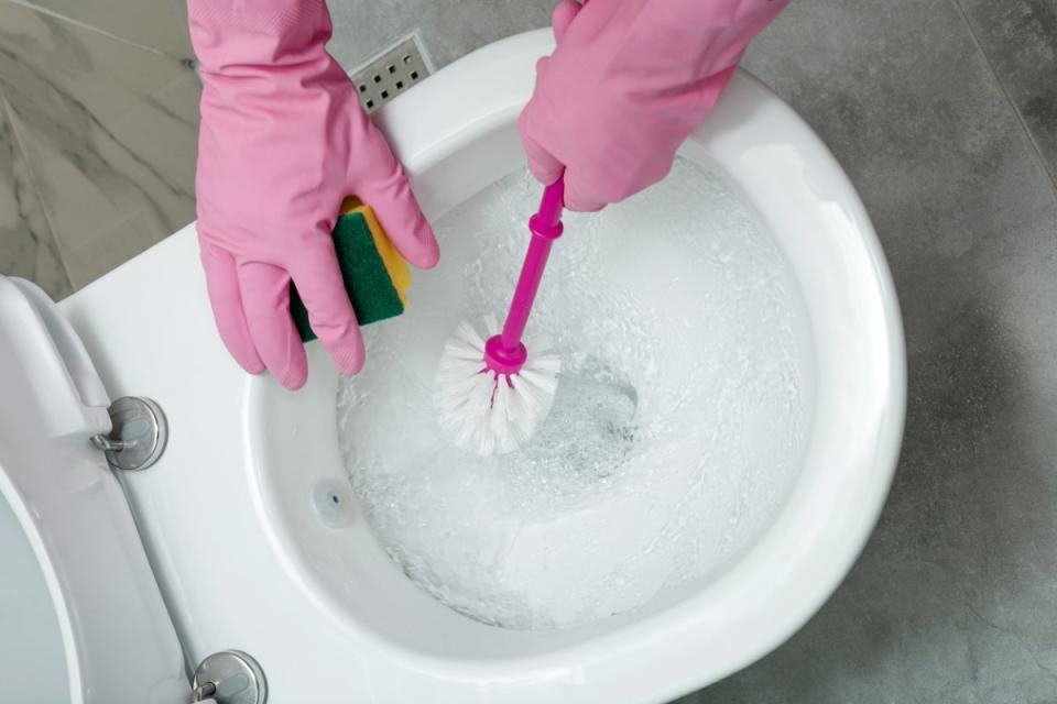 Person wearing rubber gloves cleaning toilet with a sponge and toilet brush.