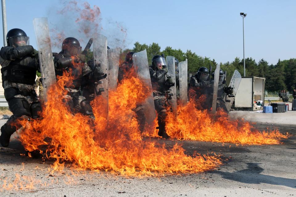 A photo of multiple soldiers standing in line in full riot gear with fire around their feet.