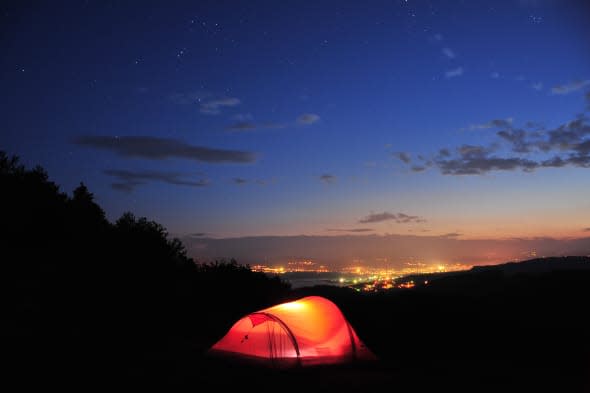 Camping at night. Lighted tent under the stars in mountains, with city lights in background, Bulgaria.