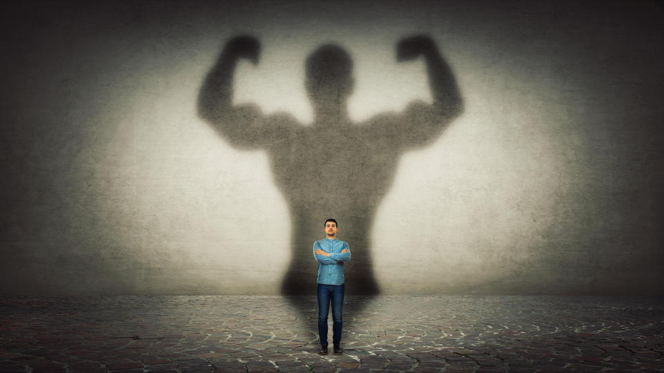 A thin man stands with his arms crossed in front of a huge, muscular shadow behind him that's three times his size.