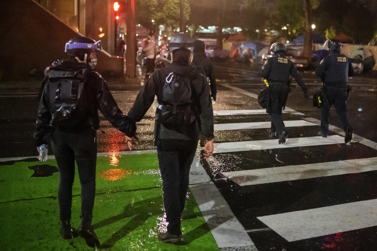 Image: Two protesters hold hands while walking near Portland police officers during an Indigenous Peoples Day of Rage protest (Nathan Howard / Getty Images)