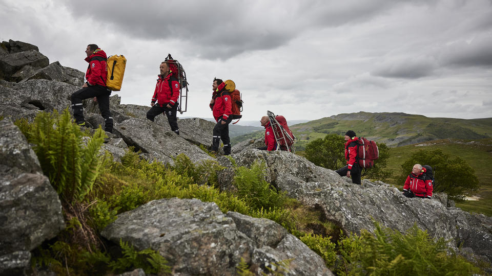 A mountain rescue team in bright red jackets walk across a rocky mountainside.