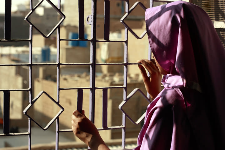 Photo shows a woman looking out a window with bars