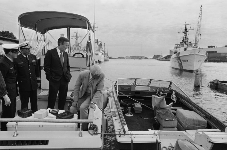 (Original Caption) Miami, Florida: Standing on a boat confiscated from drug smugglers, Vice President George Bush looks over at bales of marijuana in another boat as containers of cocaine were placed on a cooler in front of him for inspection. Admiral Richard Cueroni (L) and Cpt. John Walton (R) showed Vice President Bush what had been achieved in "Operation Hat Trick." Vice President Bush participated in awards ceremonies to various law enforcement agencies involved in the search for drug smugglers.