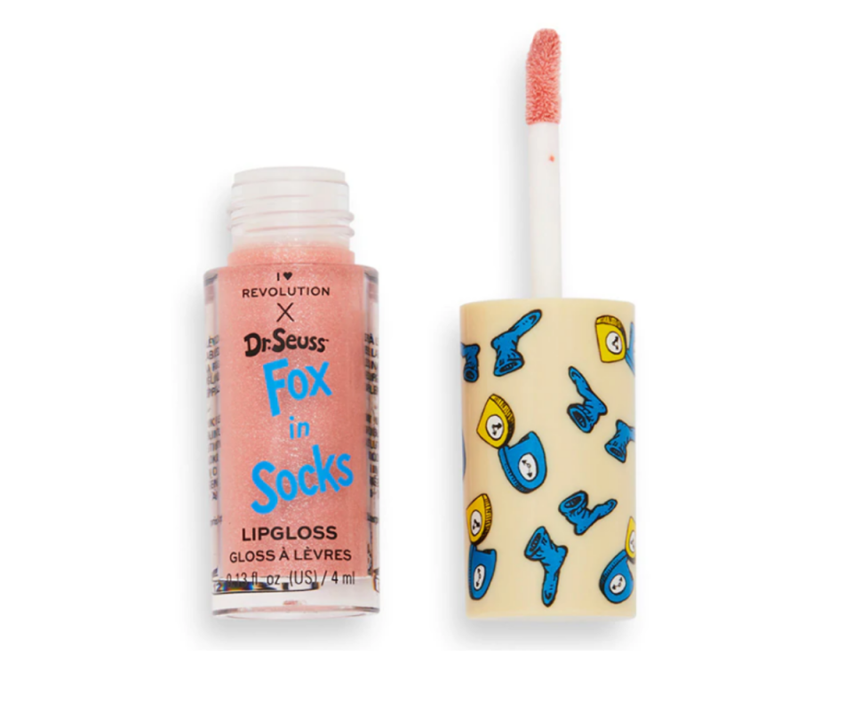 A peach-coloured lip gloss sits on the left against a white background with the lid off and to the right standing upside with the doe foot applicator facing up. Fox in Socks is written in blue on the front of the clear lip gloss bottle with tblue and yellow clocks patterned on a beige background on the lid. 