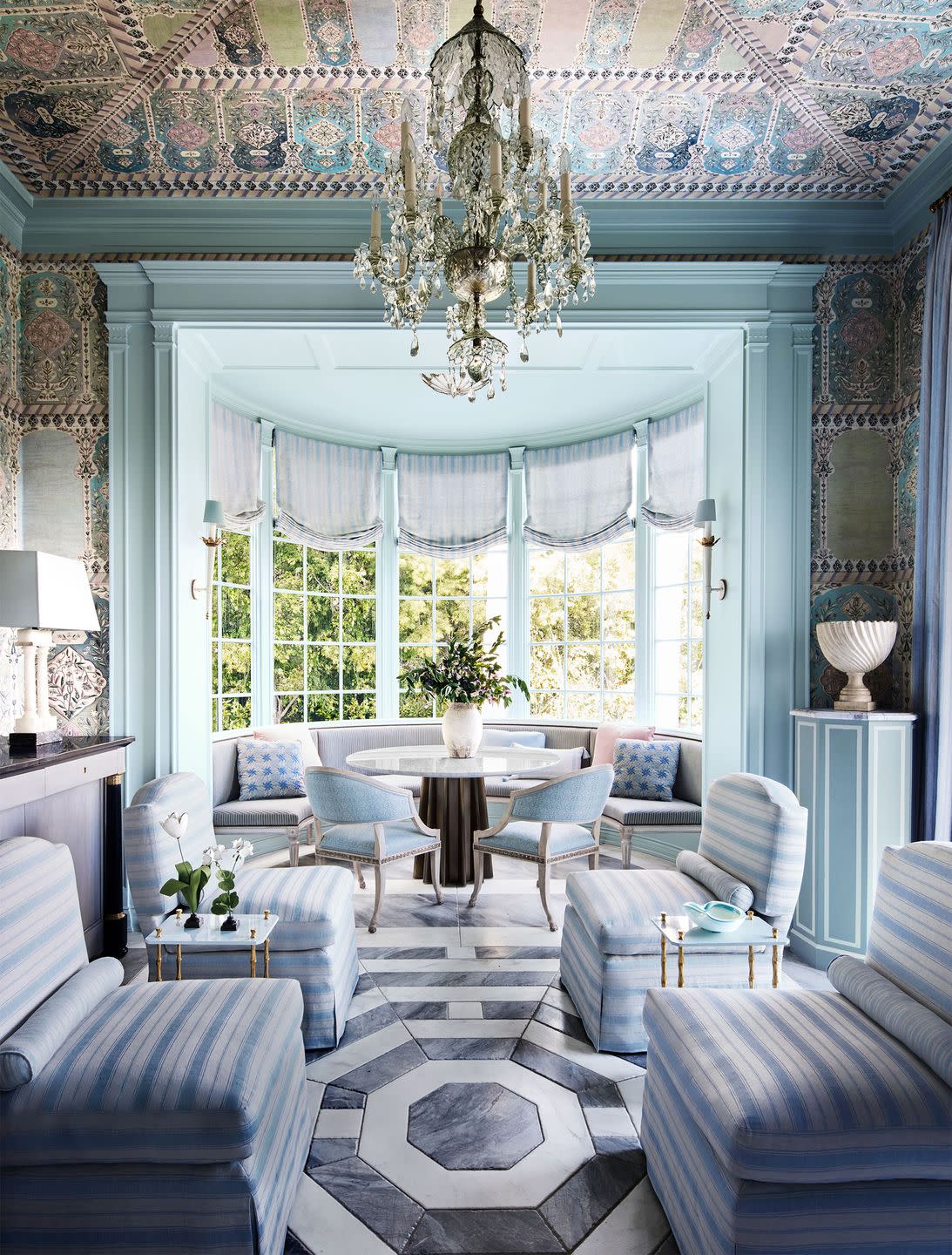 in a tea room is a bay window with a banquette and roman shades, a round table with two chairs, four blue and white striped slipper chairs, walls and ceiling covered in ottoman tent pattern, octagonal tiled floor