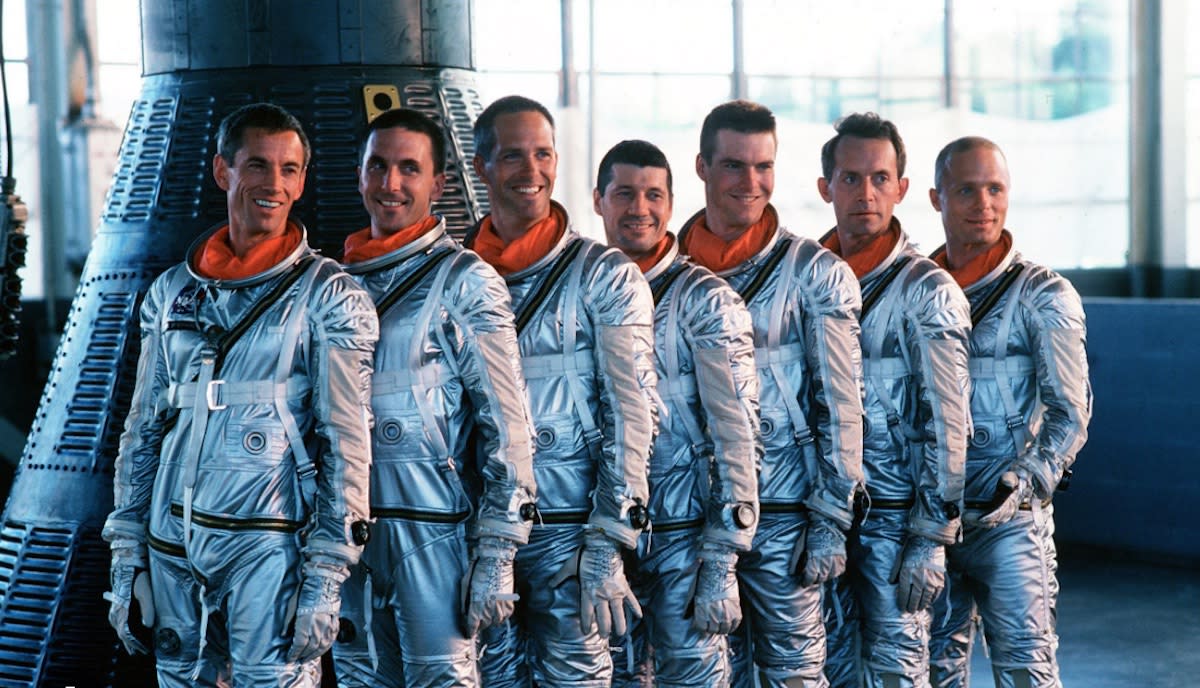  Seven men in silver spacesuits stand beside a cone-shaped spacecraft. 