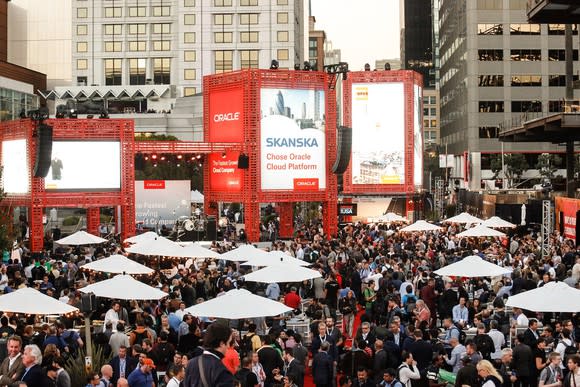 Large crowd in urban setting with elevated boards showing Oracle logo and promotional materials.