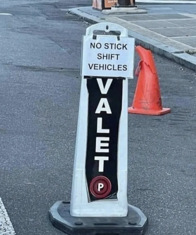 Sign on a street reads "No stick shift vehicles. VALET" with a red parking symbol below. Orange traffic cone on the sidewalk is visible in the background
