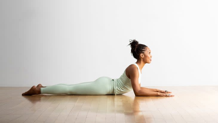 Black woman wears sea green yoga tights as she practices Sphinx pose. She is lying on a wood floor and has a white wall behind her