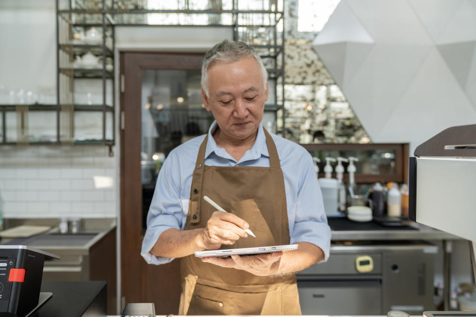 An older man wearing an apron writes on a tablet while standing in a modern kitchen