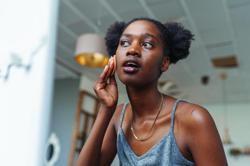 Young multiracial woman applying foundation on face with sponge while standing in front of mirror trying to hide skin problems