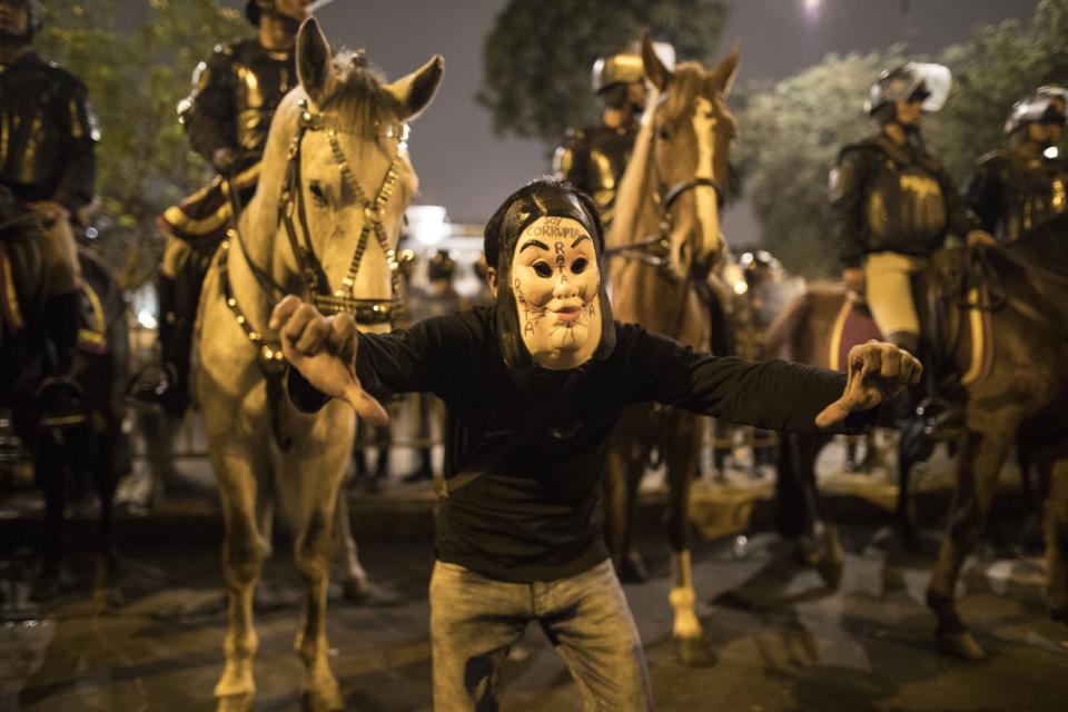 A masked supporter of President Martin Vizcarra gives a thumbs down outside Congress where horse-mounted police stand guard after the president dissolved the legislature in Lima, Peru, Monday, Sept. 30, 2019. Vizcarra dissolved congress Monday, exercising seldom used executive powers to shut down the opposition-controlled legislature that he accuses of stonewalling attempts to curb widespread corruption. (AP Photo/Rodrigo Abd)