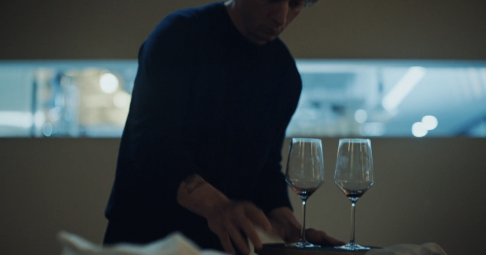 A person in a dark top handles a tray with two wine glasses in a dimly lit room, suggesting a work or dining environment