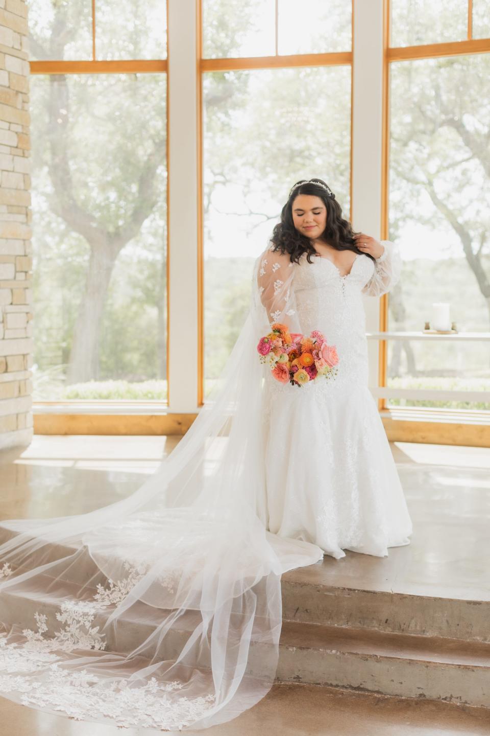 A bride looks down at her wedding dress in front of a wall of windows.