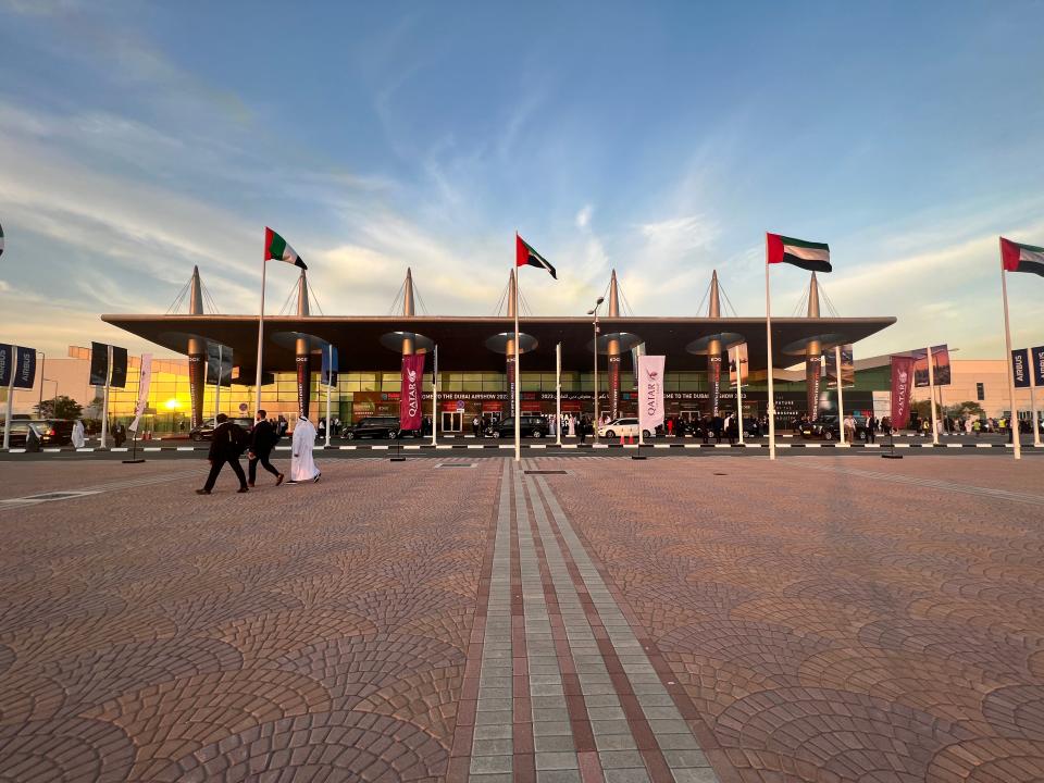 The sun sets behind the exhibition hall and entrance to the Dubai Airshow, with Qatar Airways and UAE flags in the foreground.