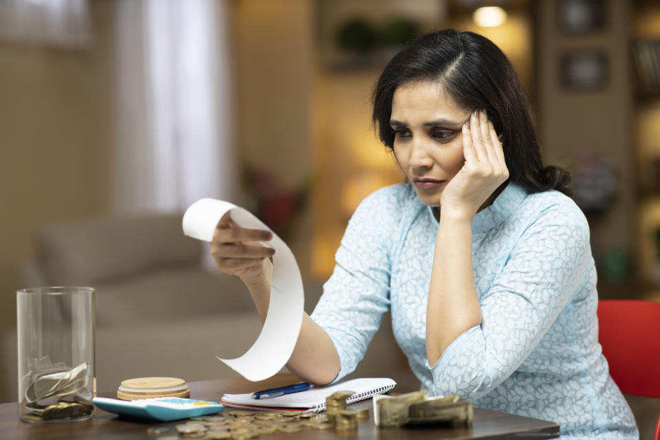 A woman reads a long receipt with a concerned expression, surrounded by coins, money, and notes at a table