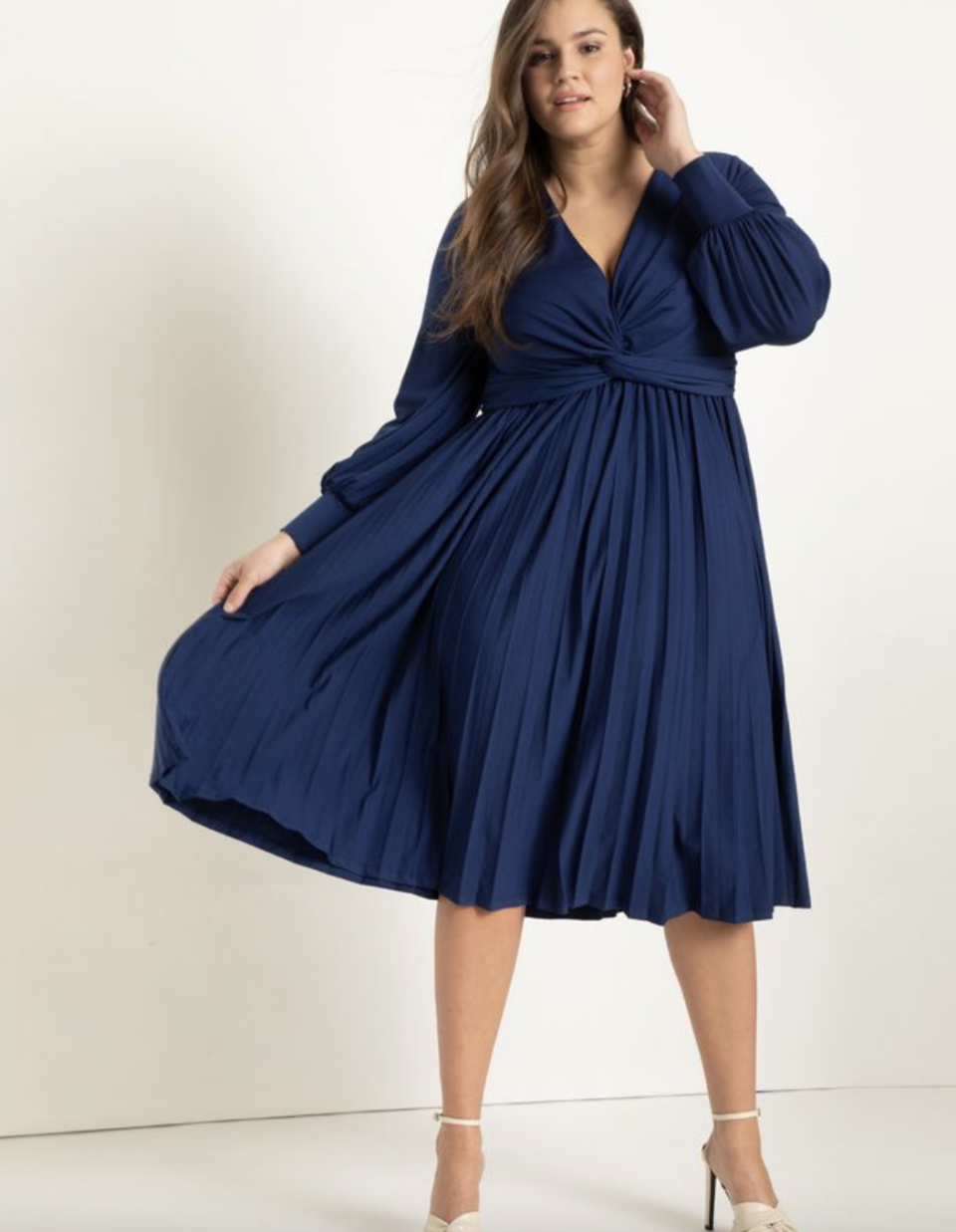plus size brunette model in long sleeve navy blue Knot Front Pleated Skirt Dress (Photo via Eloquii)
