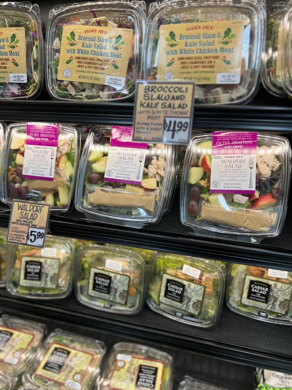 Pre-packaged salads on a store shelf, including Waldorf and Broccoli Kale Salad, priced at $5.99 and $11.99 respectively