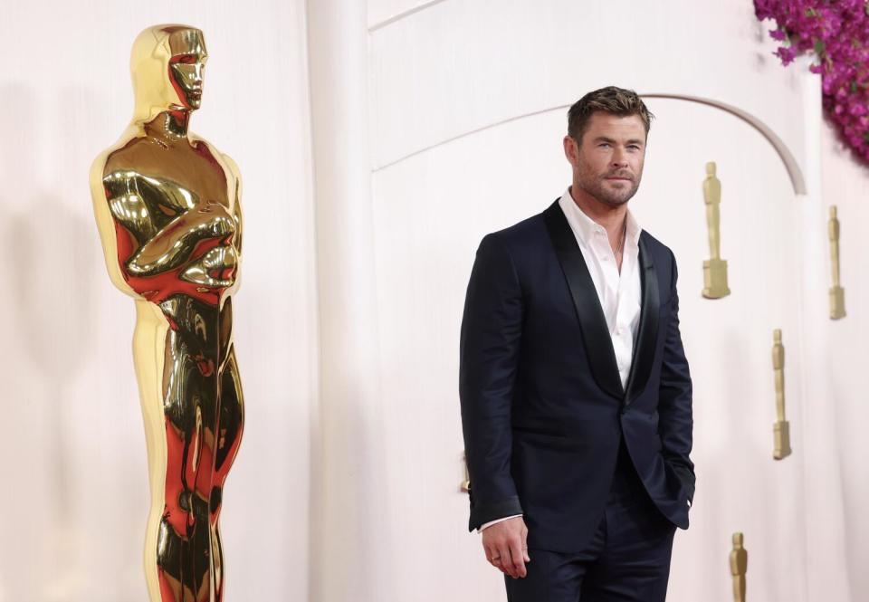 Chris Hemsworth wears a suit as he stands in front of an Oscar statue.