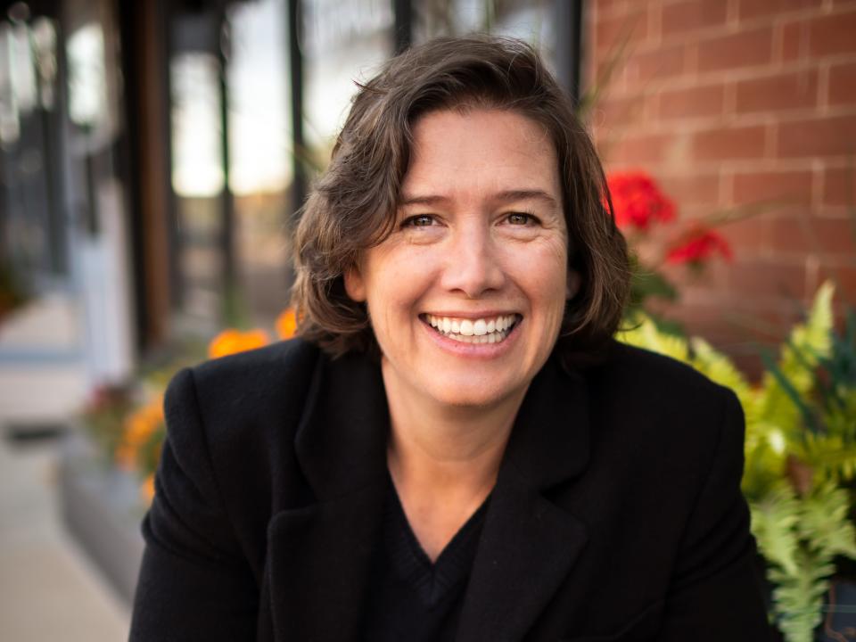 Stormy Peters is wearing a black blazer, black blouse, here hands are clasped on a black table.