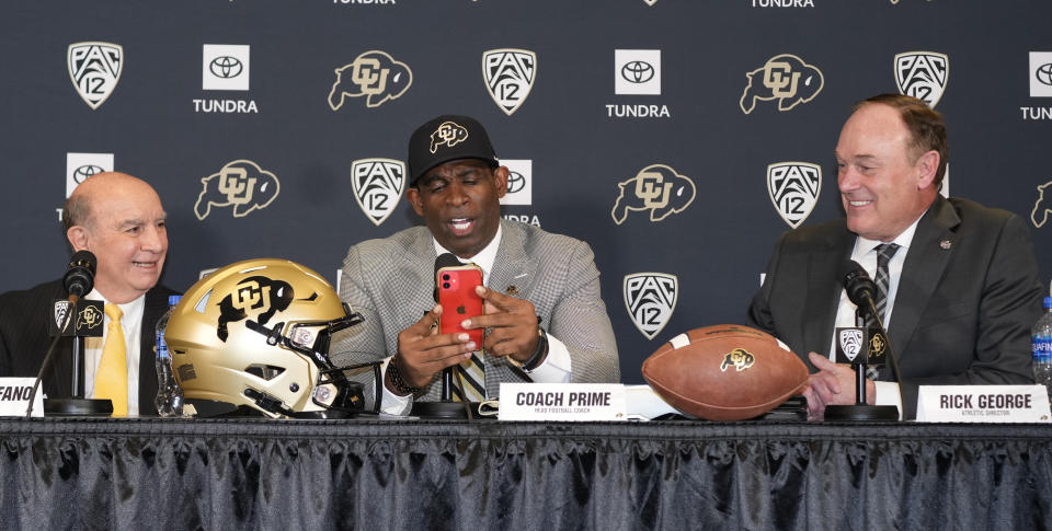 Deion Sanders, center, uses his mobile telephone to illustrate how Rick George, right, Colorado athletic director, kept in touch while courting Sanders to become the new head football coach at Colorado during a news conference Sunday, Dec. 4, 2022, in Boulder, Colo. Colorado chancellor Phil DeStefano, left, looks on. Sanders left Jackson State University after three seasons at the helm of the school's football team. (AP Photo/David Zalubowski)