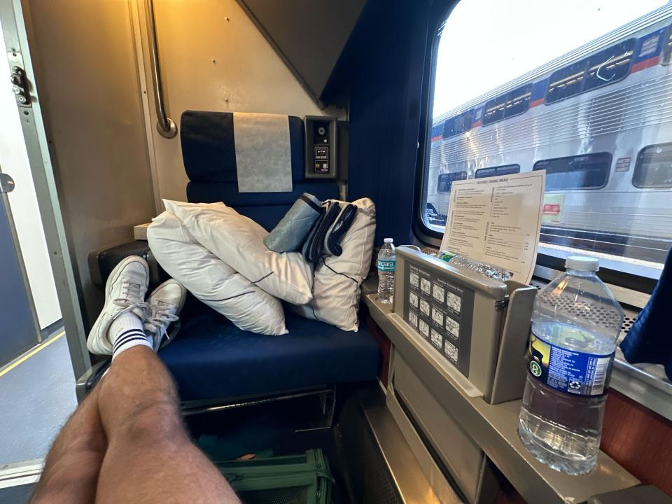 Sean's legs stretched out onto the second seat in his bedroom on an Amtrak train. Another train can be seen out the window.