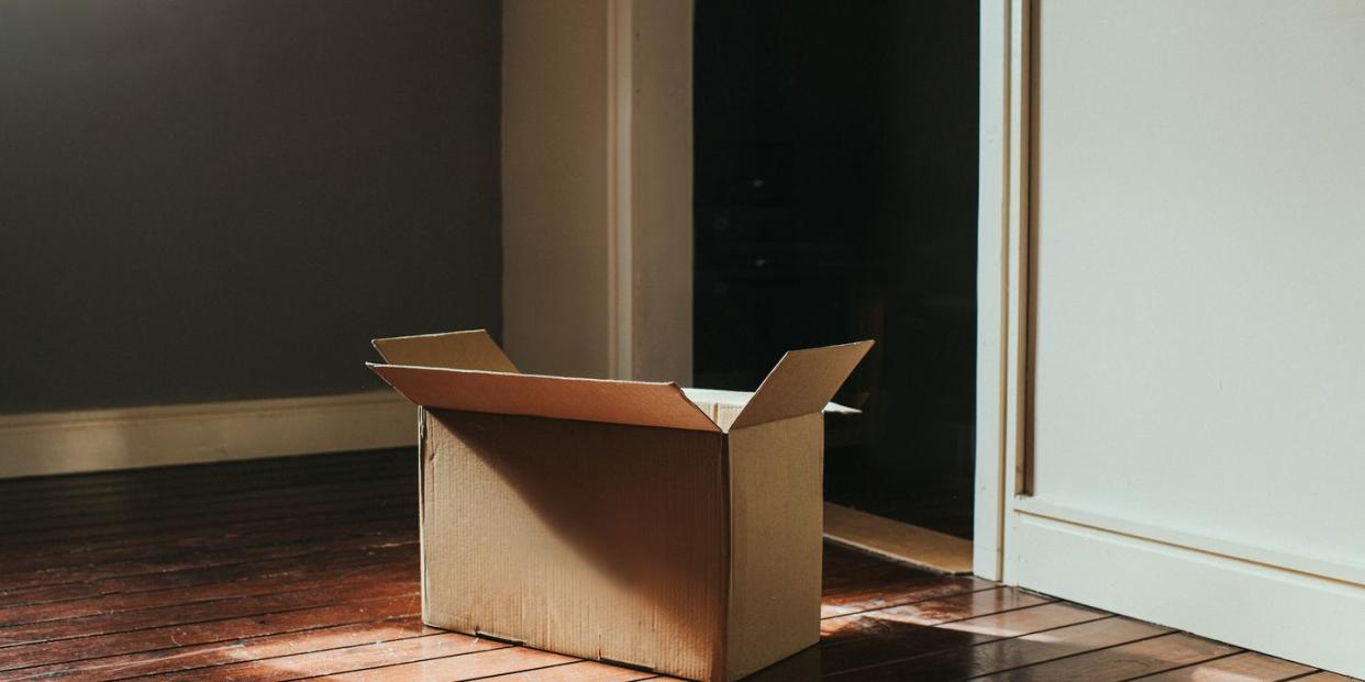 a single, plain, cardboard box sits on a dark wooden floor in a domestic room sun shines through the window creating flares and shadows room is empty and sparse wall provides a space for copy