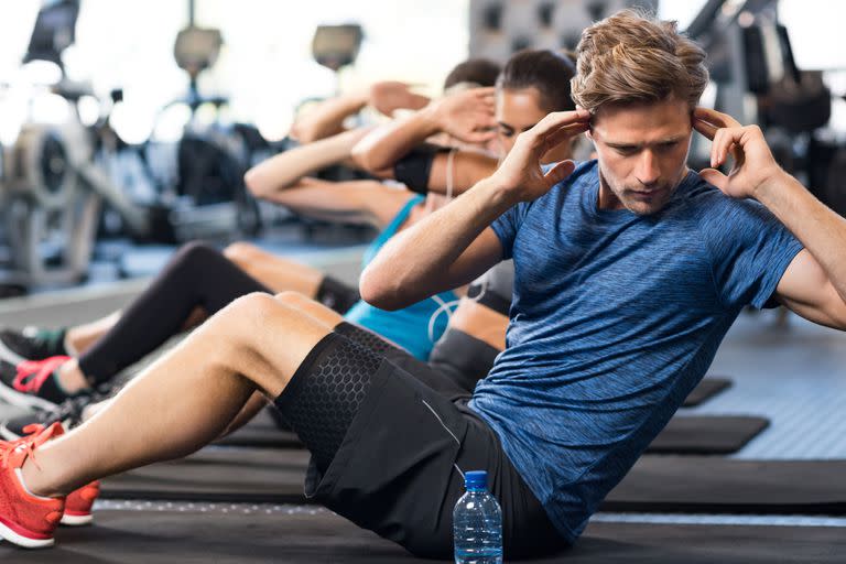 Muscular guy doing sit ups at gym with other people in background. Young athlete doing stomach workout in modern gym. Handsome fit man doing crunches at gym.