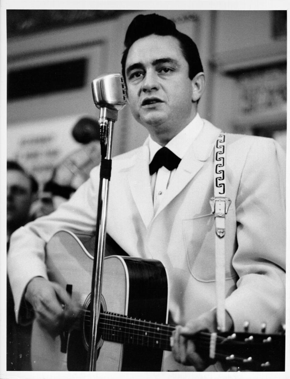 johnny cash playing a guitar and looking toward an audience behind a standing microphone