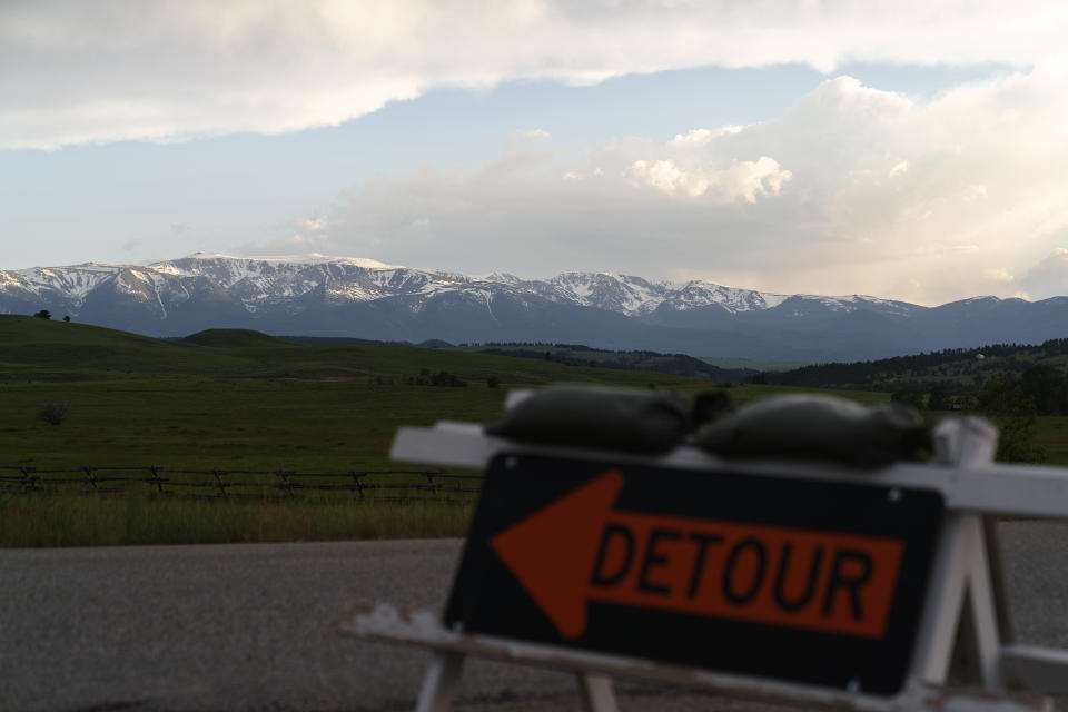 Snow capped mountains stand in the background as a detour sign directs traffic off a damaged road from severe flooding in Fishtail, Mont., Friday, June 17, 2022. (AP Photo/David Goldman)