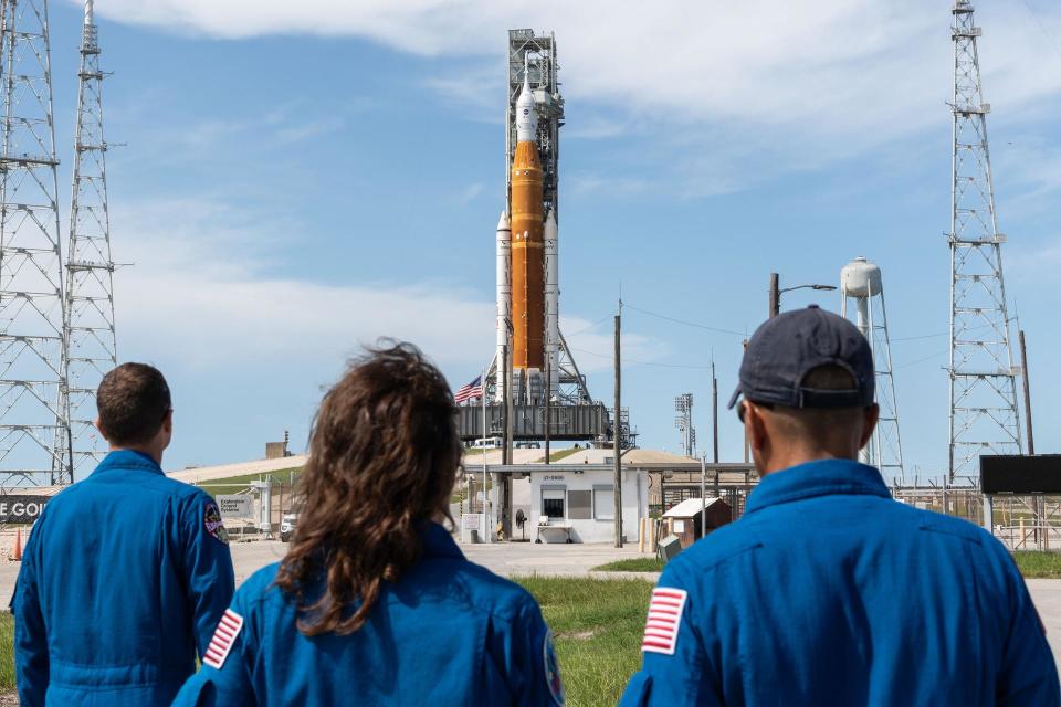 three people in blue flight suits look at a giant orange rocket on a launch pad